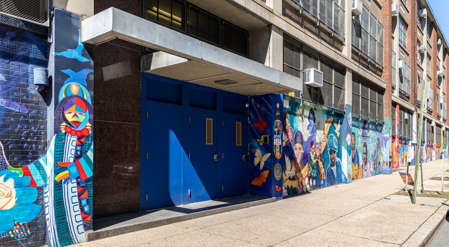 Blue entrance doors at Kenderton Elementary School surrounded by a colorful mural featuring people, butterflies and cultural imagery.